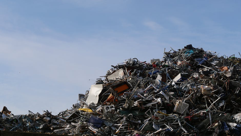 A large pile of assorted scrap metal and discarded electronic equipment dominates the image, situated outdoors against a backdrop of a clear blue sky. The heap contains various materials, including tw