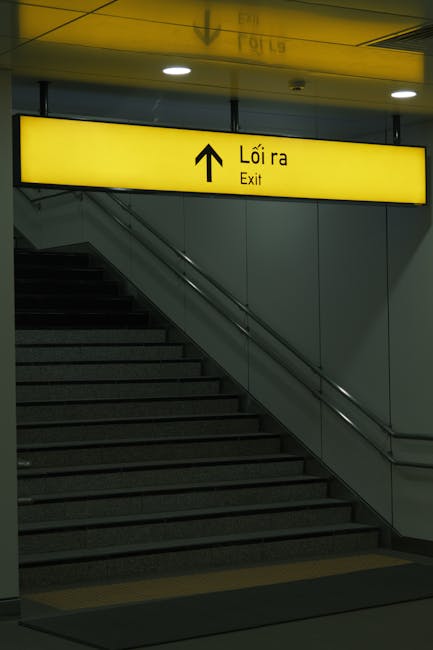 A set of indoor stairs leading upwards with dark gray, textured steps and metal handrails on both sides. Above the staircase, a large rectangular yellow sign with black text and an arrow pointing stra